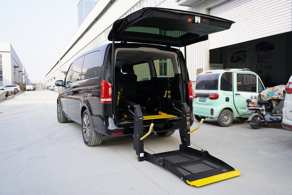 A black van with its rear hatch open, revealing a wheelchair lift mechanism positioned at the back, on an outdoor paved area adjacent to a building with a large roller shutter door. The wheelchair lift, constructed from black metal with yellow safety features, extends down to the ground, ready for loading or unloading. Inside the van, visible are cardboard boxes, wrapped furniture, and packing materials, indicating a home relocation process involving furniture transport and packing. Nearby, a small green electric vehicle and a scooter are parked, with additional vehicles visible down the street. The scene captures the preparation for a professional removals service, with the lift facilitating access for mobility-challenged individuals or heavy furniture, supporting efficient and safe loading and unloading during the move. The environment is well-lit with natural daylight, emphasizing the logistical aspects of the home's moving process handled by Man with Van Bromley as part of their removals service offerings, including narrow access solutions.