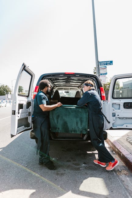 Two men dressed in casual clothing and safety goggles are engaged in a home relocation task, carefully lifting and loading a large, wrapped piece of furniture into the open cargo area of a white van parked on a street. The van is positioned near a sidewalk with a curb, and the background includes street signs, utility poles, and other vehicles, suggesting an urban environment. The interior of the van is visible, showing an empty space prepared for furniture transport, while the men use their hands to guide the item securely inside, reflecting the loading process involved in professional removals. Man with Van Bromley, a company specializing in removals, is providing a service that requires careful handling of household furniture during local moving and packing operations, with attention to space constraints and safety during the loading procedure.