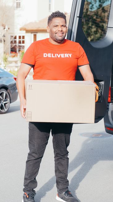A man with a beard and short curly hair, dressed in a bright orange T-shirt with the word 'DELIVERY' printed on the front, is standing outdoors next to a black vehicle with an open rear door. He is holding a large, sealed cardboard box, which appears to be part of a home relocation or furniture transport process. Behind him, residential buildings and parked cars are visible on a sunny day, indicating an urban or suburban street scene. The man is smiling slightly and appears to be preparing for or engaged in a packing and moving task, consistent with professional removals services provided by Man with Van Bromley. The scene illustrates the loading process involved in house removals, where moving staff handle boxes and furniture with care using appropriate packaging materials and equipment, such as straps or trolleys, to facilitate transportation from a property to a vehicle.