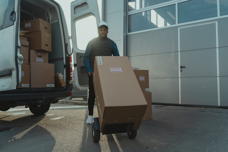 A man wearing a white cap, navy blue jacket, and black trousers is loading a large cardboard box onto a black hand truck outside a white van parked on a paved driveway, with the van's sliding door open revealing more packed cardboard boxes inside. The boxes are sealed with packing tape marked with the logo of Man with Van Bromley, and some are stacked inside the vehicle, indicating a home relocation or furniture transport process. The scene takes place in daylight, with a modern building structure visible in the background, and the exterior environment suggests a residential or commercial property in the Bromley South BR1 area. The presence of packing materials, the loading activity, and the open van door reflect the logistics involved in moving services, including packing, loading, and transportation for a house removal. Man with Van Bromley specializes in removals, assisting clients with efficient packing and home relocation tasks.