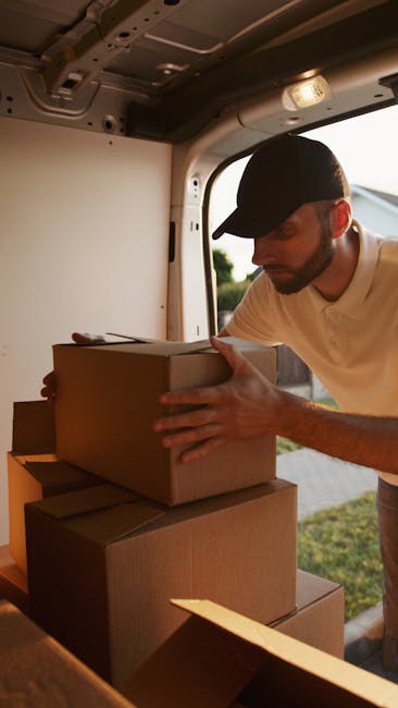 A man with a beard and short curly hair, dressed in a bright orange T-shirt with the word 'DELIVERY' printed on the front, is standing outdoors next to a black vehicle with an open rear door. He is holding a large, sealed cardboard box, which appears to be part of a home relocation or furniture transport process. Behind him, residential buildings and parked cars are visible on a sunny day, indicating an urban or suburban street scene. The man is smiling slightly and appears to be preparing for or engaged in a packing and moving task, consistent with professional removals services provided by Man with Van Bromley. The scene illustrates the loading process involved in house removals, where moving staff handle boxes and furniture with care using appropriate packaging materials and equipment, such as straps or trolleys, to facilitate transportation from a property to a vehicle.