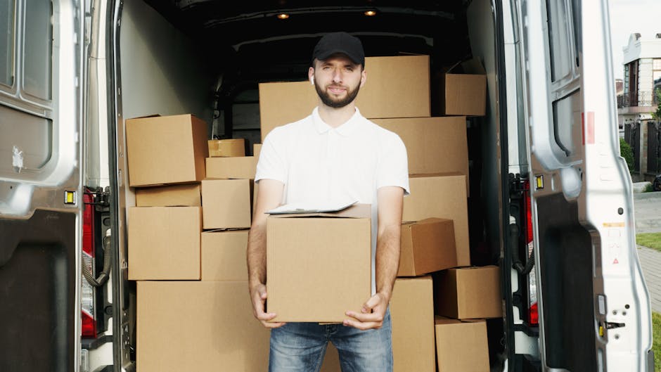 A young male mover with light brown, wavy hair, wearing a black bandana and a navy blue shirt, is sitting inside a white delivery van with the sliding door open. He is smiling and giving a thumbs-up g
